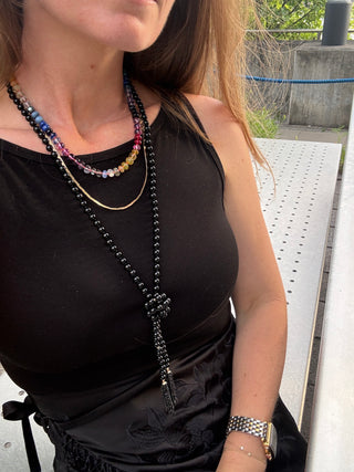 A close-up lifestyle shot of a woman wearing a long black beaded lariat necklace layered over a black top, with other layered necklaces and a gold watch visible.