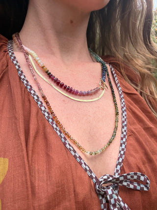 Close-up of a person wearing multiple necklaces with a checkered ribbon tie on a rust-colored garment.