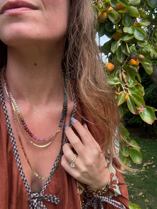 Woman wearing multiple necklaces with a blurred background of greenery and fruits.