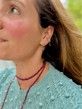 Close-up of a person wearing gold jellyfish earrings and a ruby beaded necklace, with a blurred natural background.