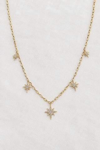 A delicate gold chain necklace featuring five small starburst charms, with a larger central starburst pendant, displayed on a textured white fabric background.